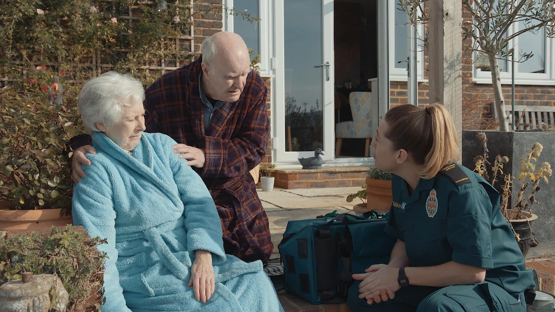 An old gent is talking to a female paramedic while holding a suspected stroke female patient 